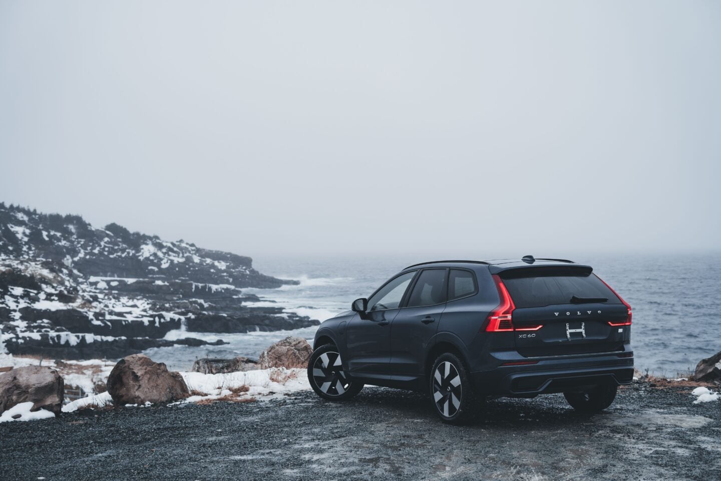 Dark blue Volvo XC60 luxury SUV viewed from the rear three-quarter angle, parked beside a dramatic snow-covered rocky coastline, with its distinctive vertical LED taillights illuminated and the ocean in the background.