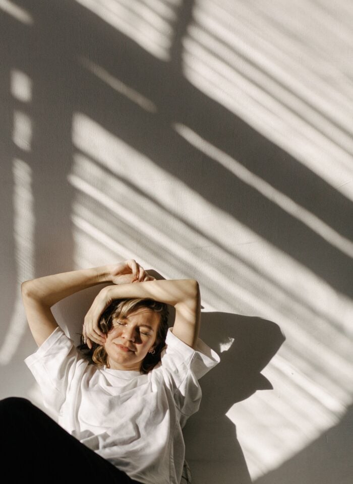 Woman relaxing with her eyes closed and arms raised above her head, enjoying a peaceful moment in natural sunlight.