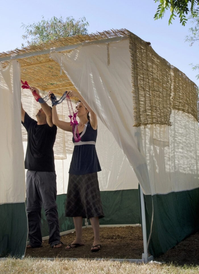 Two adults decorating a traditional outdoor sukkah in preparation for the Jewish festival of Sukkot. A man wearing a black T-shirt and grey trousers and a woman wearing a dark sleeveless top and long skirt stand inside the temporary structure, reaching upwards to hang colourful pink, red, and blue garlands from the ceiling. The sukkah features a roof made from natural reeds or branches, woven reed panels, and white fabric walls with dark green lower sections. Sunlight filters through the roof, casting soft shadows across the interior. The structure stands in a peaceful garden surrounded by leafy green trees, shrubs, cacti, and dry grass, beneath a clear blue sky. The scene captures the traditional Sukkot activity of decorating the sukkah for the Jewish harvest festival.