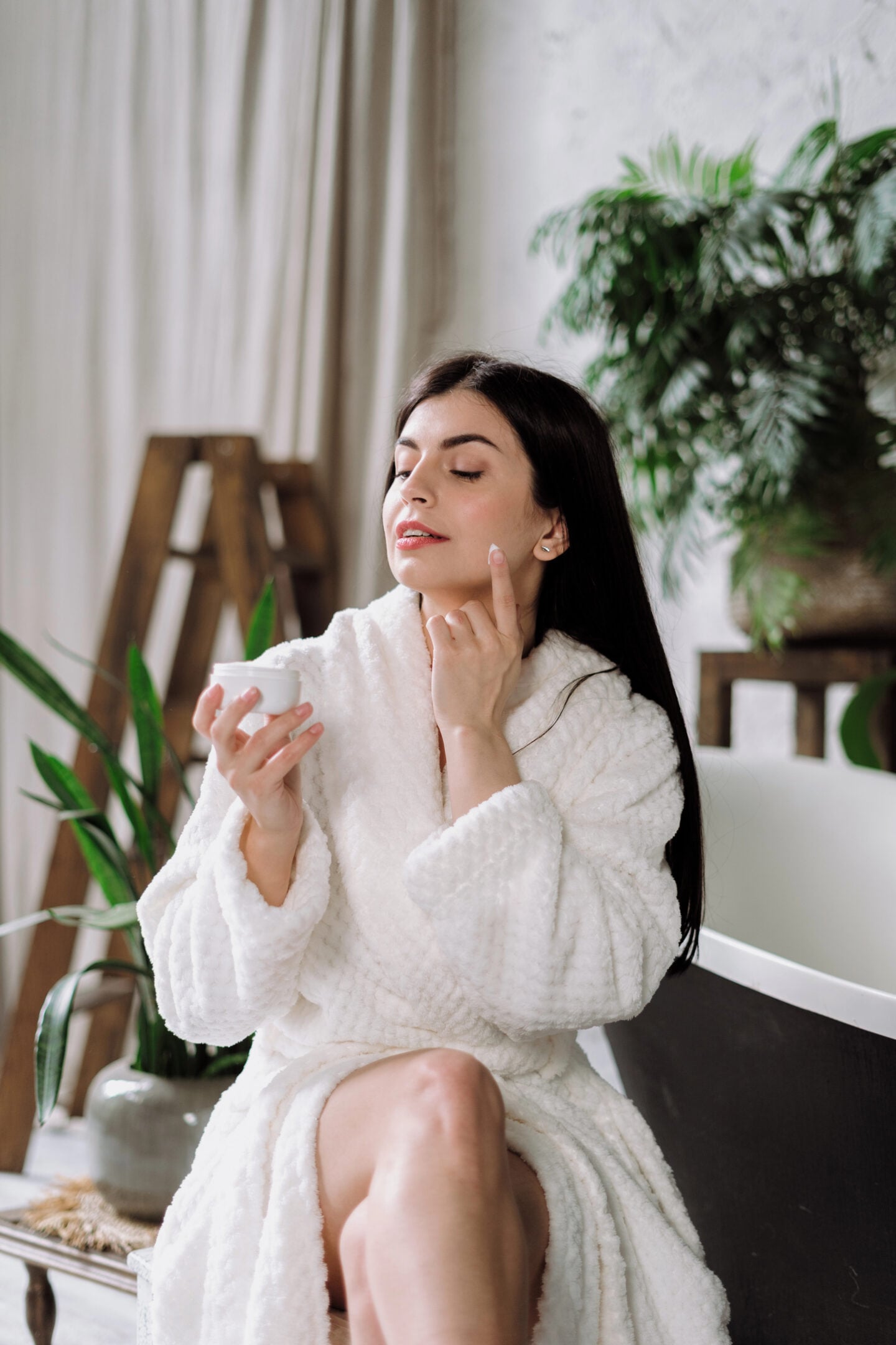 A woman with long, straight dark brown hair wearing a fluffy white bathrobe sits in a bright, elegant room while applying moisturizing face cream as part of her daily skincare routine. She holds a small white jar of skincare cream in one hand and gently dabs a small amount of white moisturizer onto her cheek with the index finger of her other hand. Her eyes are softly closed, and she has a relaxed expression, neatly shaped eyebrows, subtle makeup, and small stud earrings. The cozy interior features soft cream-colored curtains, a wooden decorative ladder, leafy green houseplants in ceramic pots, and a modern black-and-white bench. Natural daylight illuminates the scene, creating a peaceful, spa-like atmosphere that reflects self-care, facial hydration, and an at-home beauty routine for glowing skin.