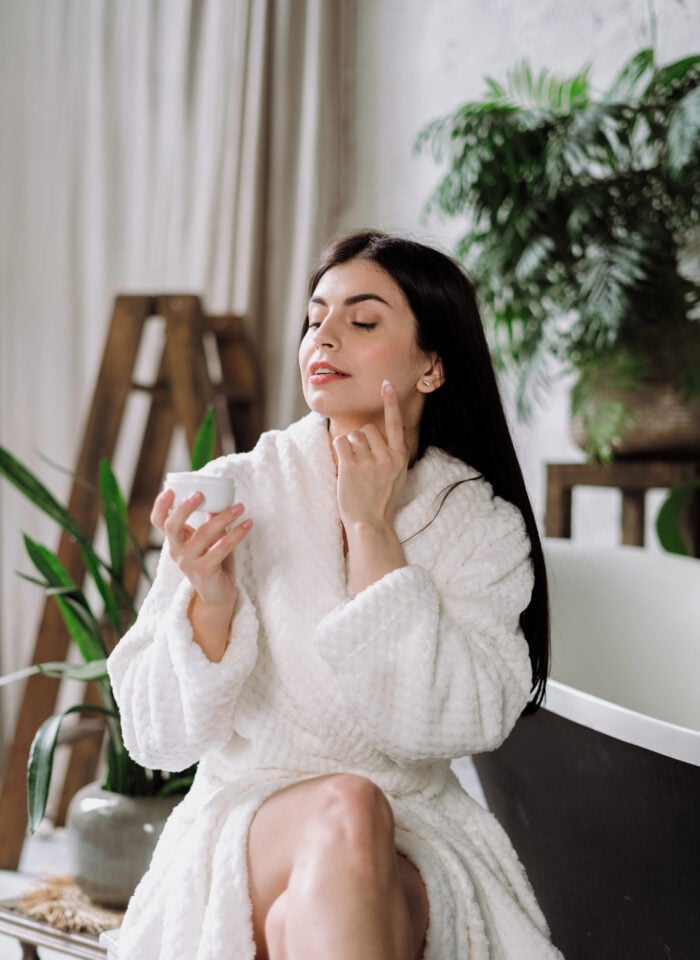 A woman with long, straight dark brown hair wearing a fluffy white bathrobe sits in a bright, elegant room while applying moisturizing face cream as part of her daily skincare routine. She holds a small white jar of skincare cream in one hand and gently dabs a small amount of white moisturizer onto her cheek with the index finger of her other hand. Her eyes are softly closed, and she has a relaxed expression, neatly shaped eyebrows, subtle makeup, and small stud earrings. The cozy interior features soft cream-colored curtains, a wooden decorative ladder, leafy green houseplants in ceramic pots, and a modern black-and-white bench. Natural daylight illuminates the scene, creating a peaceful, spa-like atmosphere that reflects self-care, facial hydration, and an at-home beauty routine for glowing skin.