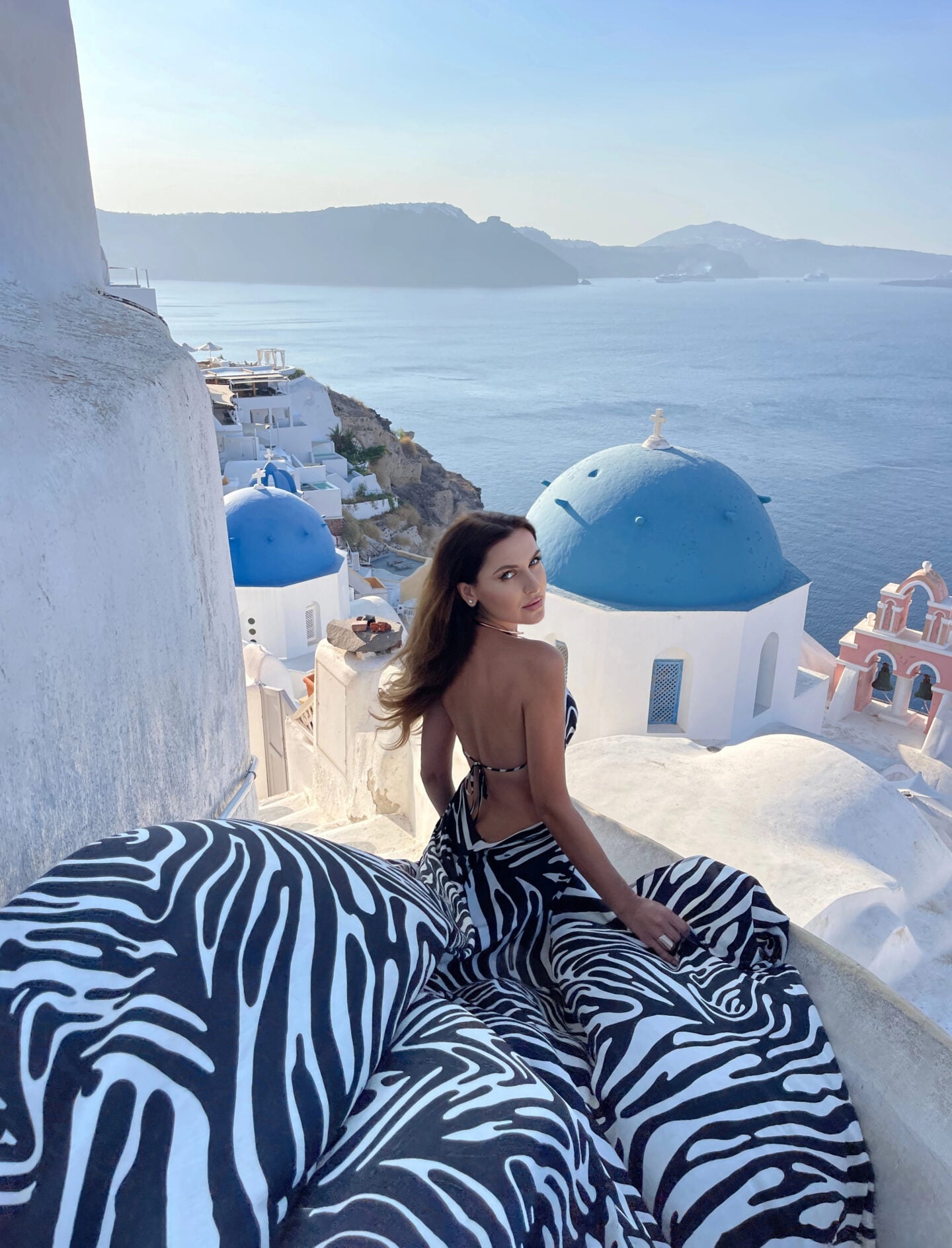Woman wearing a flowing black-and-white zebra-print backless dress overlooking the iconic blue-domed churches, whitewashed buildings, and sparkling Aegean Sea in Oia, Santorini, Greece.