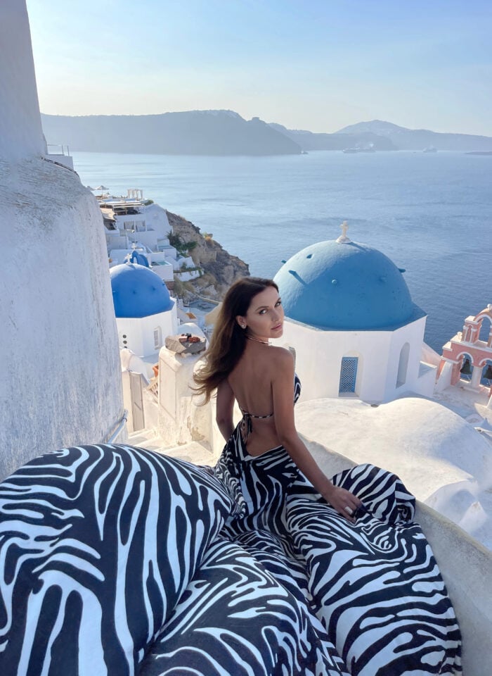 Woman wearing a flowing black-and-white zebra-print backless dress overlooking the iconic blue-domed churches, whitewashed buildings, and sparkling Aegean Sea in Oia, Santorini, Greece.