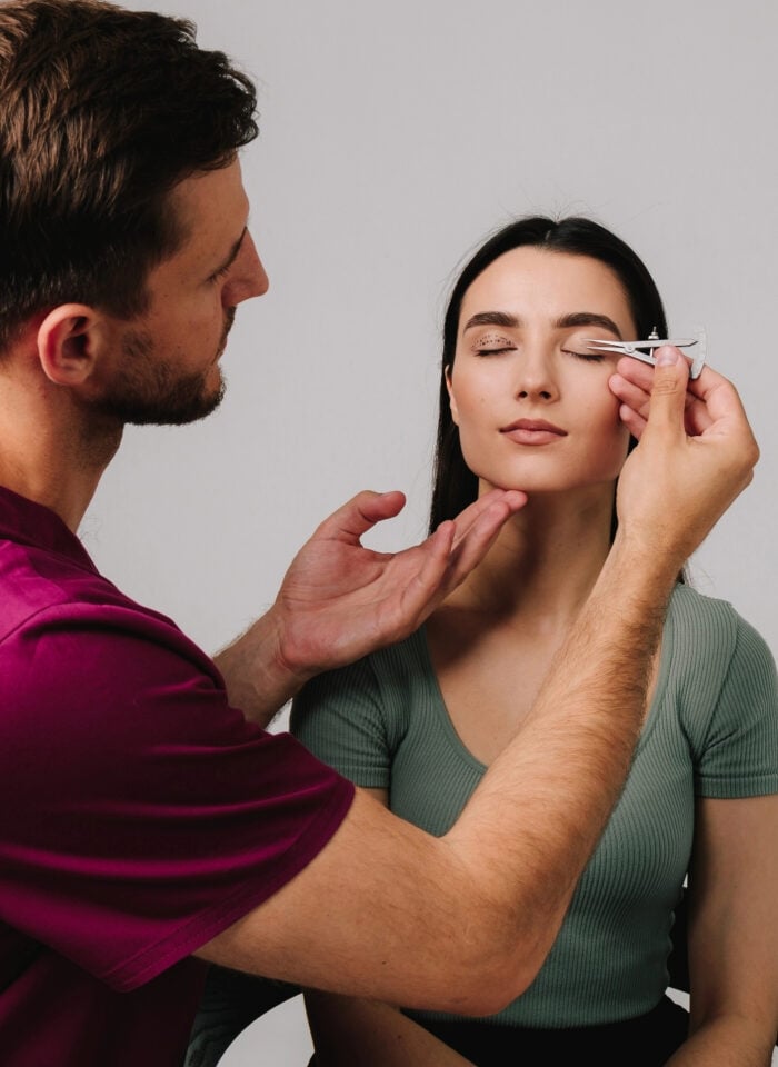Practitioner in burgundy scrubs measuring a woman’s upper eyelid with calipers, with dotted markings visible on her other eyelid.
