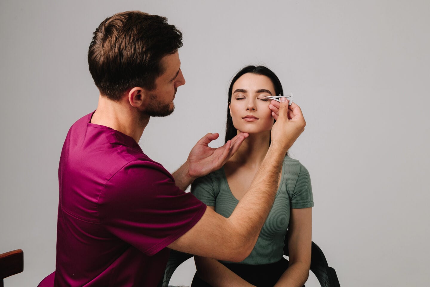 Practitioner in burgundy scrubs measuring a woman’s upper eyelid with calipers, with dotted markings visible on her other eyelid.