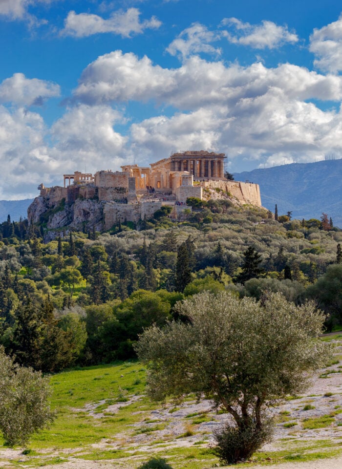 The Acropolis in Athens, Greece, with the Parthenon rising above a rocky hill, olive trees in the foreground, and white clouds across a blue sky.