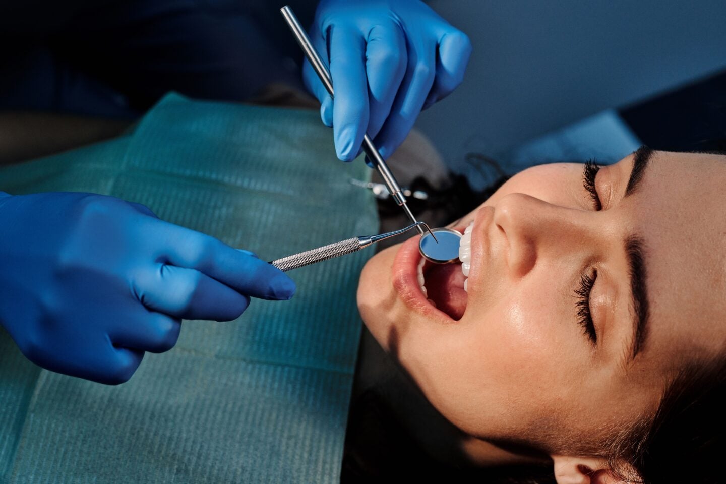 Woman undergoing a routine dental checkup, with a dentist wearing blue gloves using a dental mirror and probe to examine her teeth and gums.