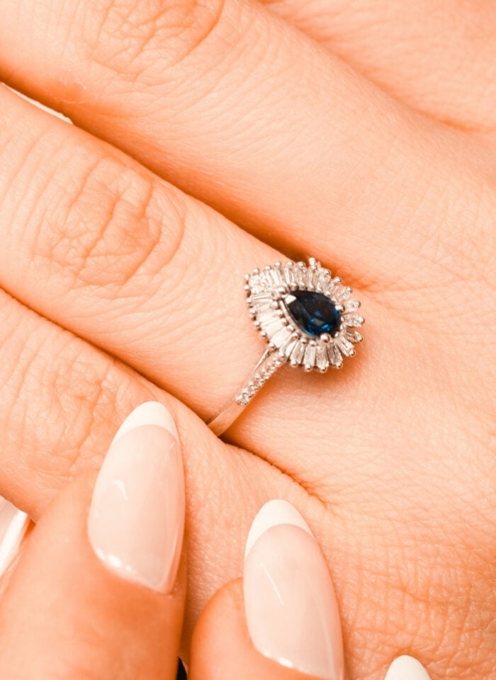 Close-up of a silver ring featuring a dark blue pear-shaped gemstone surrounded by a halo of small, clear rectangular stones. The ring is worn on a hand with long, almond-shaped nails featuring a glossy nude pink base and crisp white French tips.
