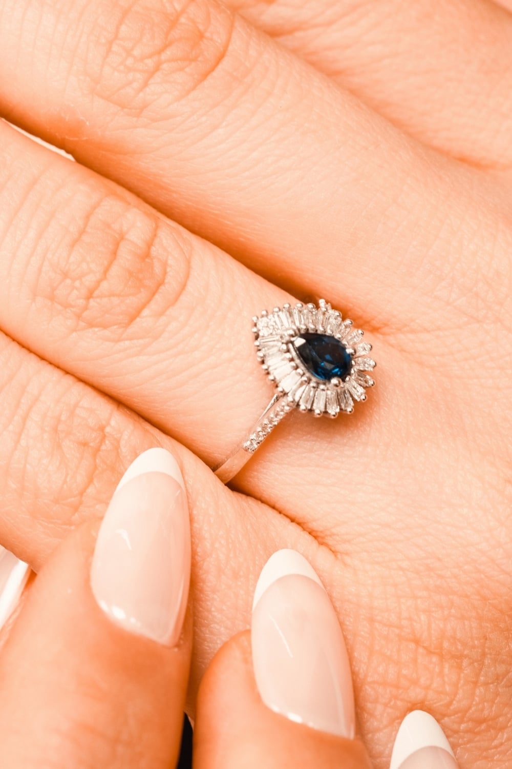 Close-up of a silver ring featuring a dark blue pear-shaped gemstone surrounded by a halo of small, clear rectangular stones. The ring is worn on a hand with long, almond-shaped nails featuring a glossy nude pink base and crisp white French tips.