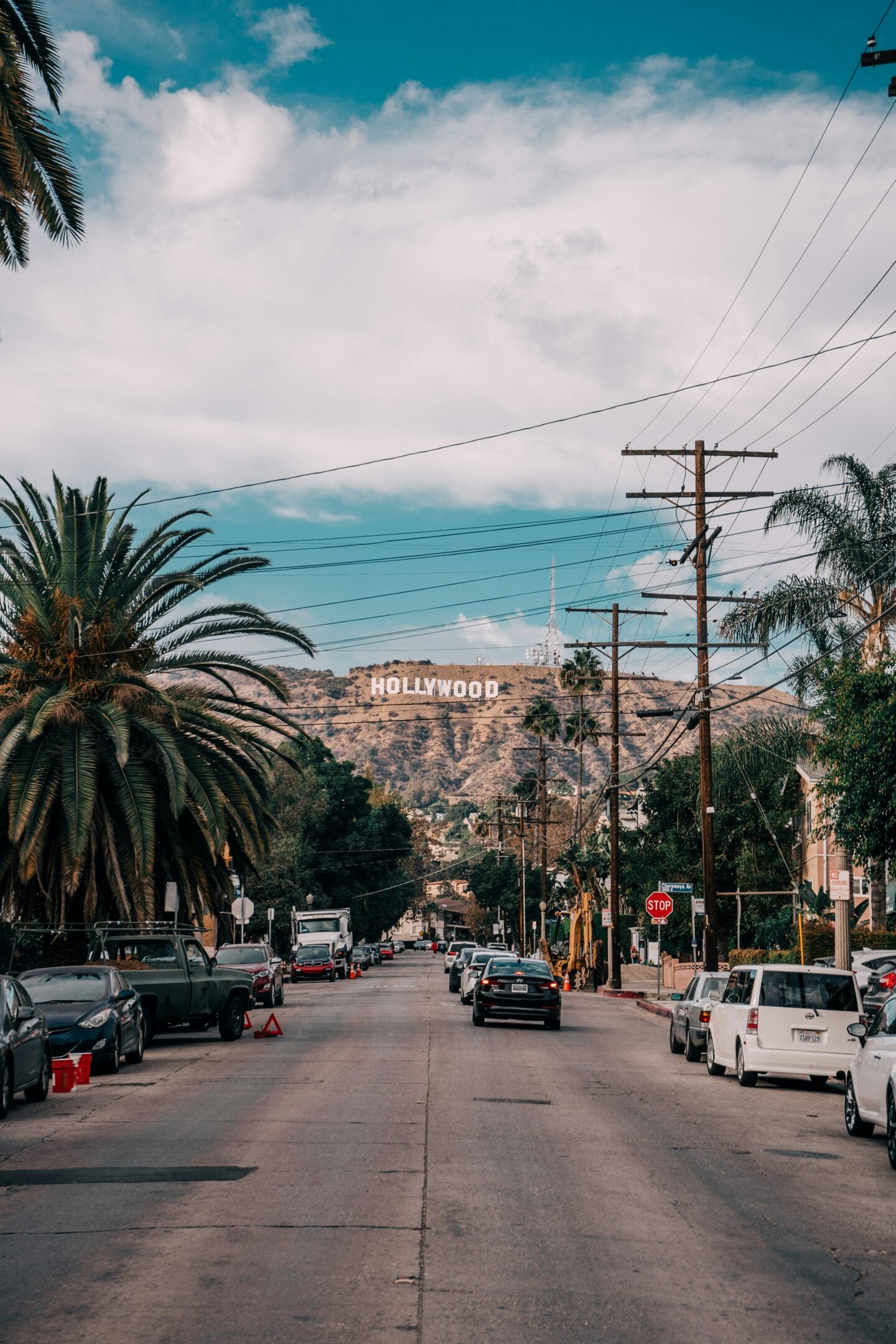 Residential street in Los Angeles with palm trees, parked cars, and utility lines leading toward the Hollywood sign on the hillside.