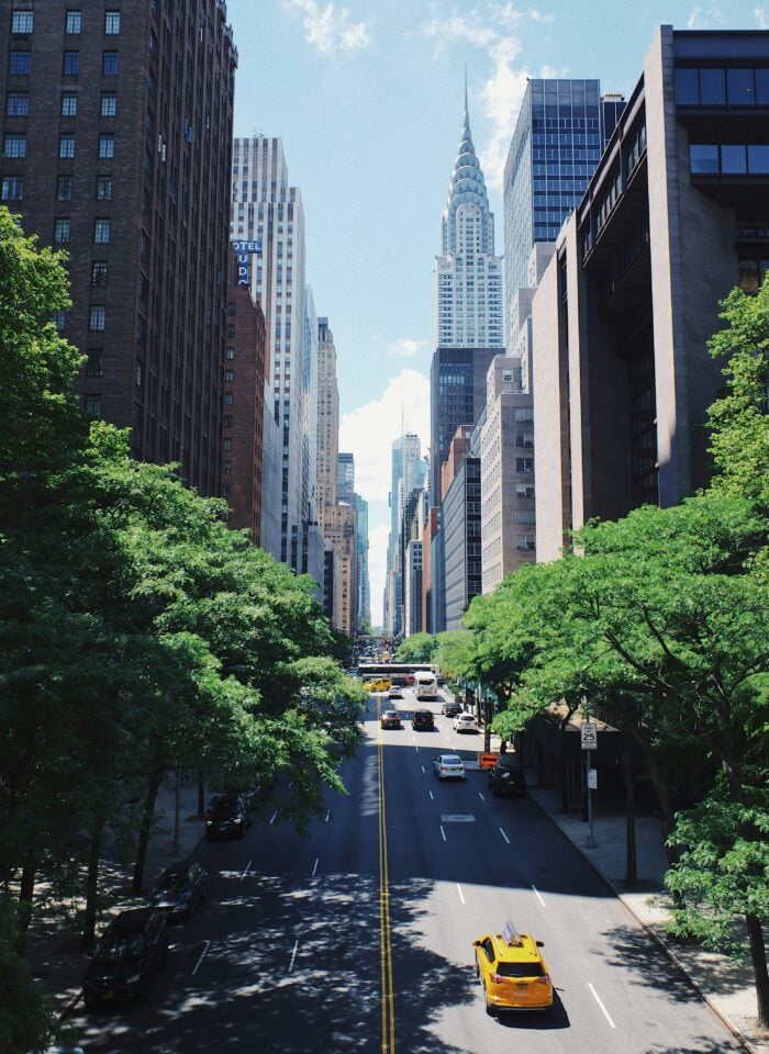 Tree-lined Manhattan street with yellow taxis and tall buildings framing the Chrysler Building beneath a blue sky.