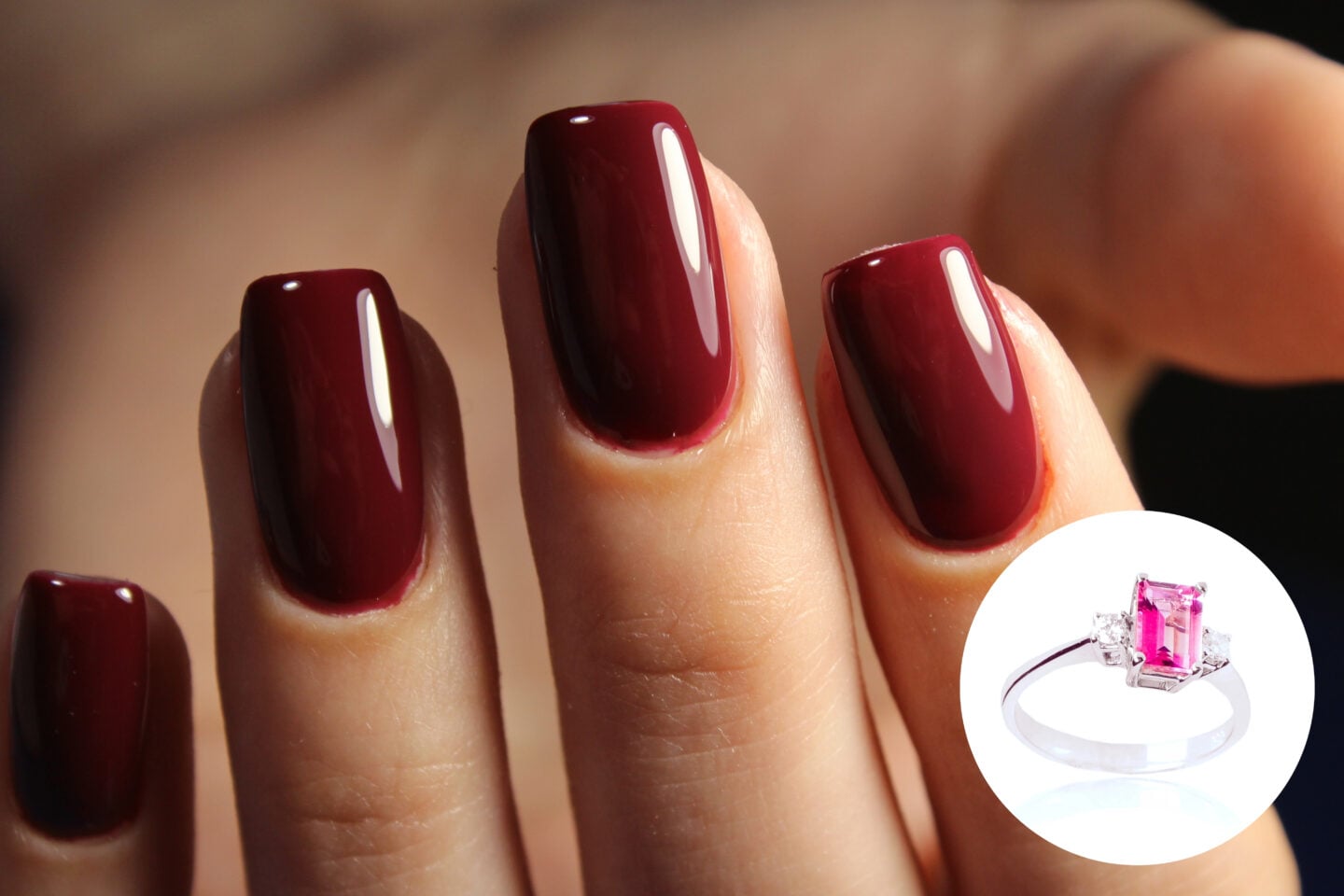 Close-up of four fingernails painted in a deep, glossy burgundy cherry red shade, with a softly squared shape and rounded corners. A circular inset in the bottom right shows a silver-coloured ring featuring a bright pink rectangular gemstone, flanked by small clear stones on either side, against a white background.